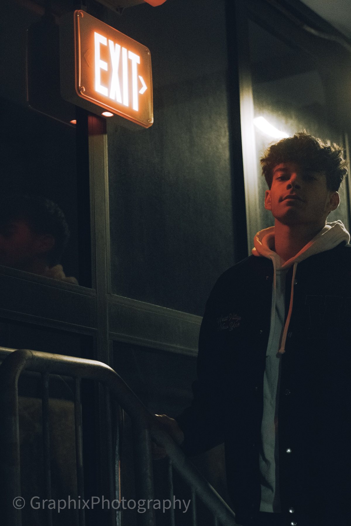 A young man on a stairway at night, lit by a glowing orange exit sign above him